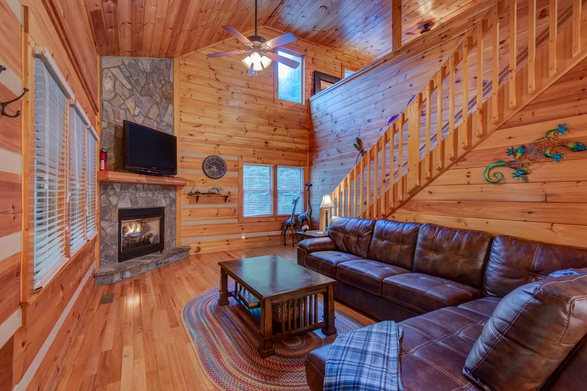A cozy living room scene centered around a stately stone fireplace, with a plush leather couch and large windows that frame views of the mountains. The wooden staircase and decorative accents enhance the warm, welcoming atmosphere of the cabin's hub.