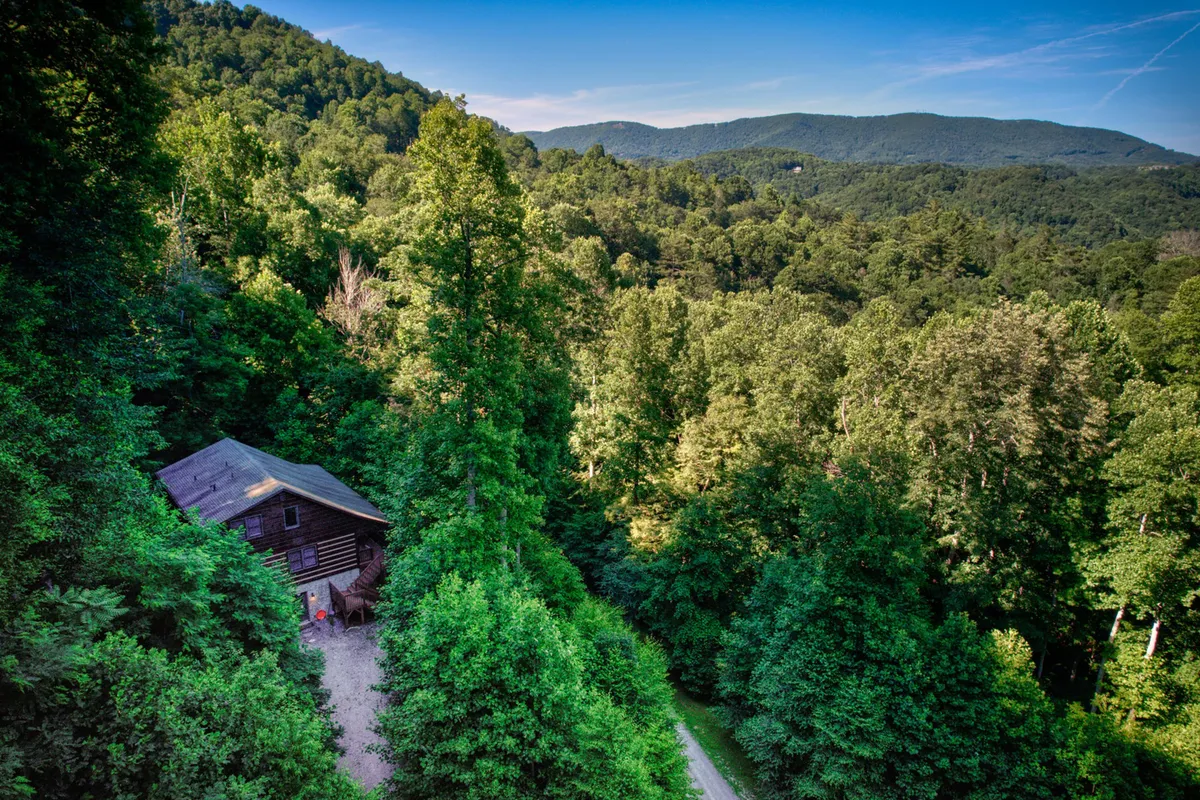 A charming log cabin is nestled among tall trees, with a mountain backdrop and a winding pathway leading up to the entrance. The setting exudes tranquility, promising both privacy and access to nature's wonders. The cabin's porch hints at a perfect place to relax outdoors, while the surrounding landscape invites exploration and adventure.