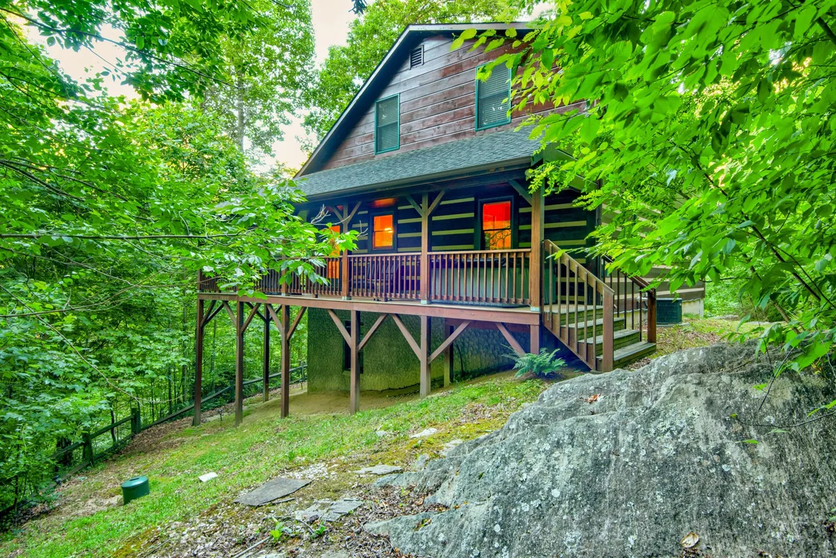 A cozy wooden deck attached to a rustic log cabin, surrounded by vibrant greenery and forest. The spacious deck offers ample room for outdoor seating and activities, with the scenic backdrop of nature providing an inviting atmosphere for relaxation and gatherings.