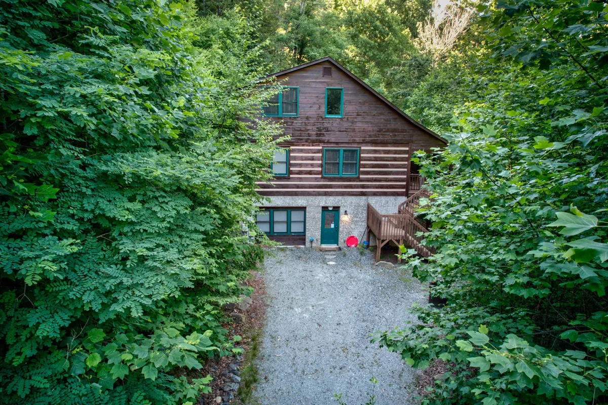 The image displays the wooden house nestled amid lush greenery, with a gravel driveway and a welcoming porch. The natural setting around the house exudes tranquility, inviting guests for outdoor relaxation or family gatherings in a peaceful environment.