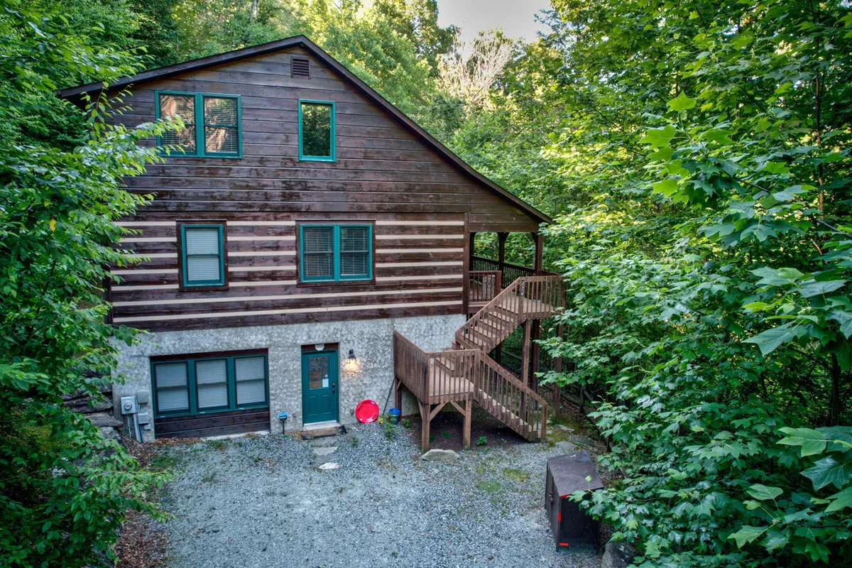 The image shows the rustic wooden exterior of a charming log cabin nestled among towering trees, with inviting green accents and a welcoming staircase leading to a spacious porch—perfect for relaxing and soaking in the natural beauty of the Blue Ridge Mountains.