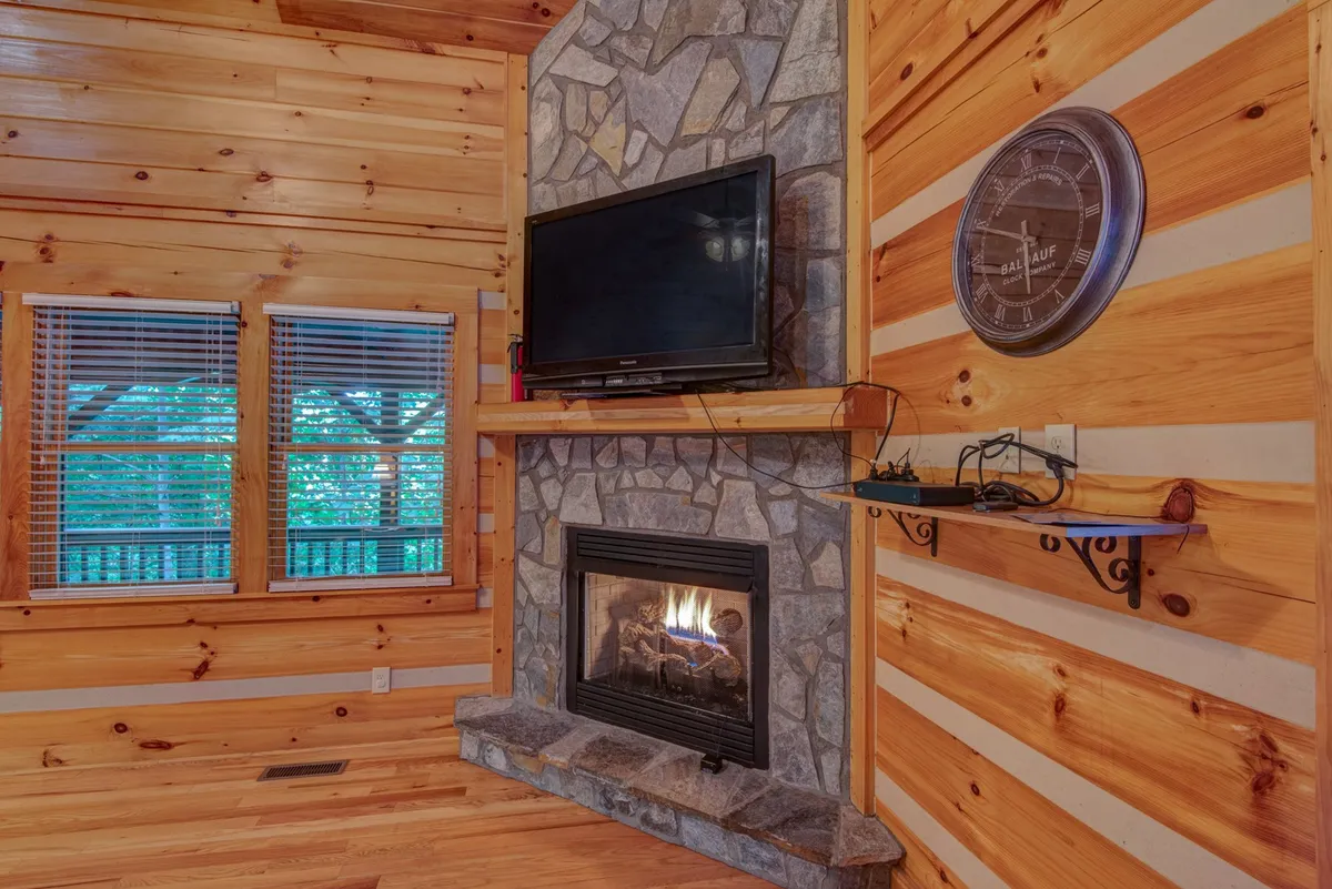 A stone fireplace anchors a welcoming living space with wooden walls and large windows. The room is designed for comfort, featuring seating that encourages gatherings and offers views of the surrounding nature through the windows.