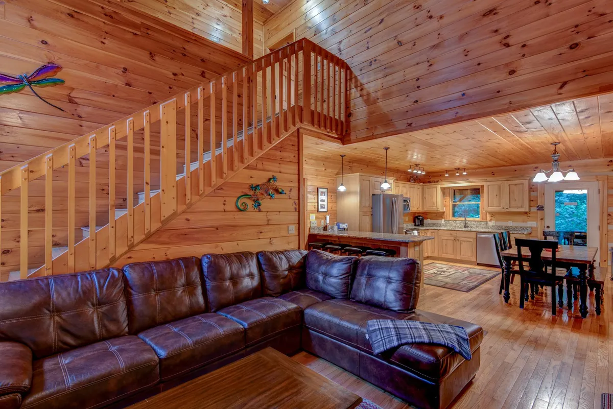 This image presents an inviting open-concept living space that seamlessly connects the living room, dining area, and kitchen. The area features a leather sectional sofa, a rustic wooden staircase, and a communal dining table, all bathed in natural light from expansive windows. The kitchen displays wooden cabinets and ample space for meal preparation and socializing.