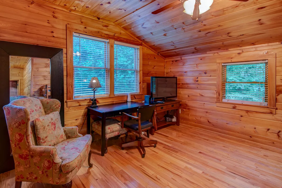 A sunlit interior featuring a floral armchair positioned near a window, with a black desk, television, and lamp creating a cozy nook. The scene captures a peaceful moment perfect for reading, working, or simply enjoying the natural light and woodland views outside.
