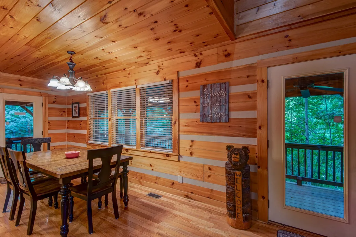 A warm, inviting dining area with a wooden table, black chairs, large windows, and a decorative bear statue. A sign on the wall and thoughtful decorative touches evoke a welcoming, familial spirit, ideal for shared meals and board games.