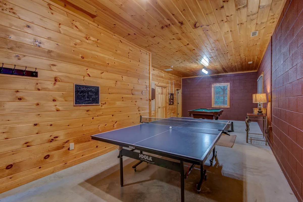 The image features a spacious, recreational interior with both a ping pong table and a pool table as prominent elements. The wall-mounted chalkboard adds a casual, competitive vibe, while a table lamp provides cozy lighting. The space is open, inviting, and clearly designed for group activities and friendly competition.