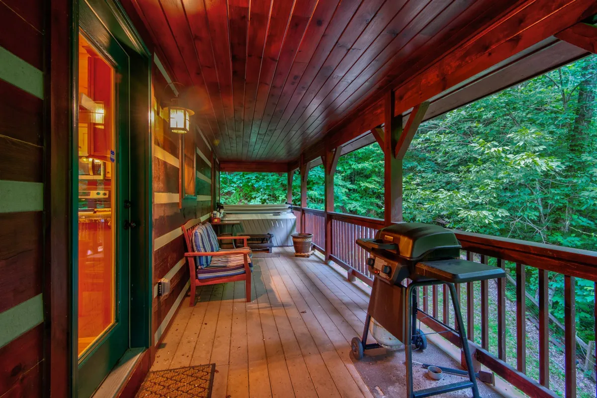 A spacious wooden deck that wraps around the log home, featuring a hot tub, gas grill, and a classic rocking chair. Lush greenery surrounds the deck, and the area is bathed in soft natural light, evoking a sense of relaxation and privacy.