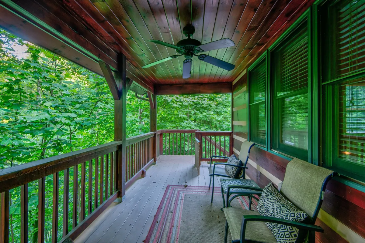 A peaceful sitting area on the porch features wooden railings, comfortable chairs, and lush green foliage in the background. The space invites guests to relax, read, or simply enjoy the sounds and sights of nature.