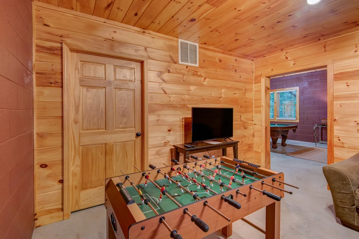 The image showcases a foosball table in the foreground and a television mounted on the wall, with a doorway leading to another room. The setup is casual and inviting, perfect for both gaming and watching movies or sports with friends and family.