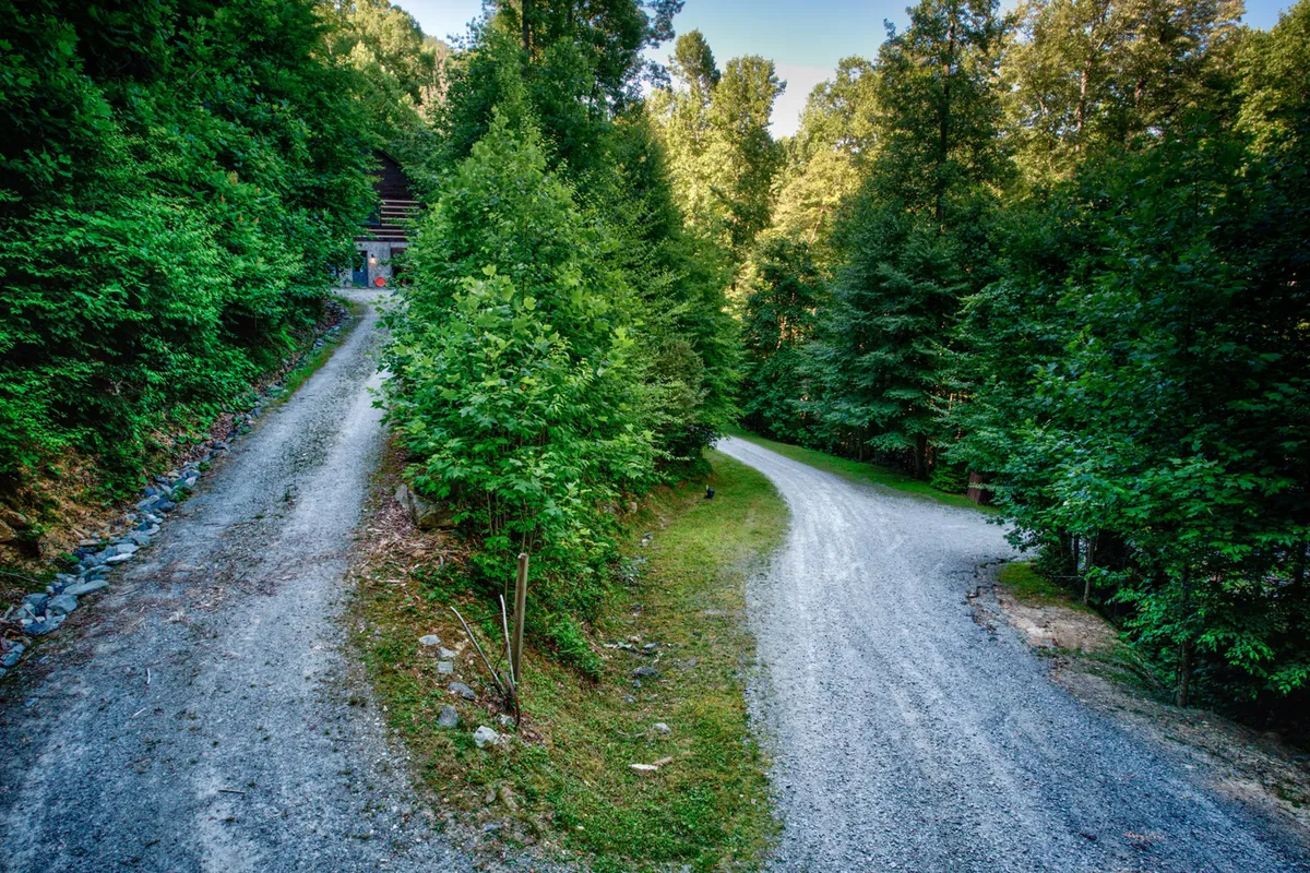 A tranquil gravel path winds through dense green foliage, dappled with sunlight. The inviting trailhead suggests opportunities for hiking, nature walks, and exploration directly from the property.